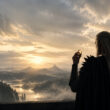 Sauron de dos, vêtu de noir, tient l'Anneau Unique devant un paysage fantastique au coucher du soleil.